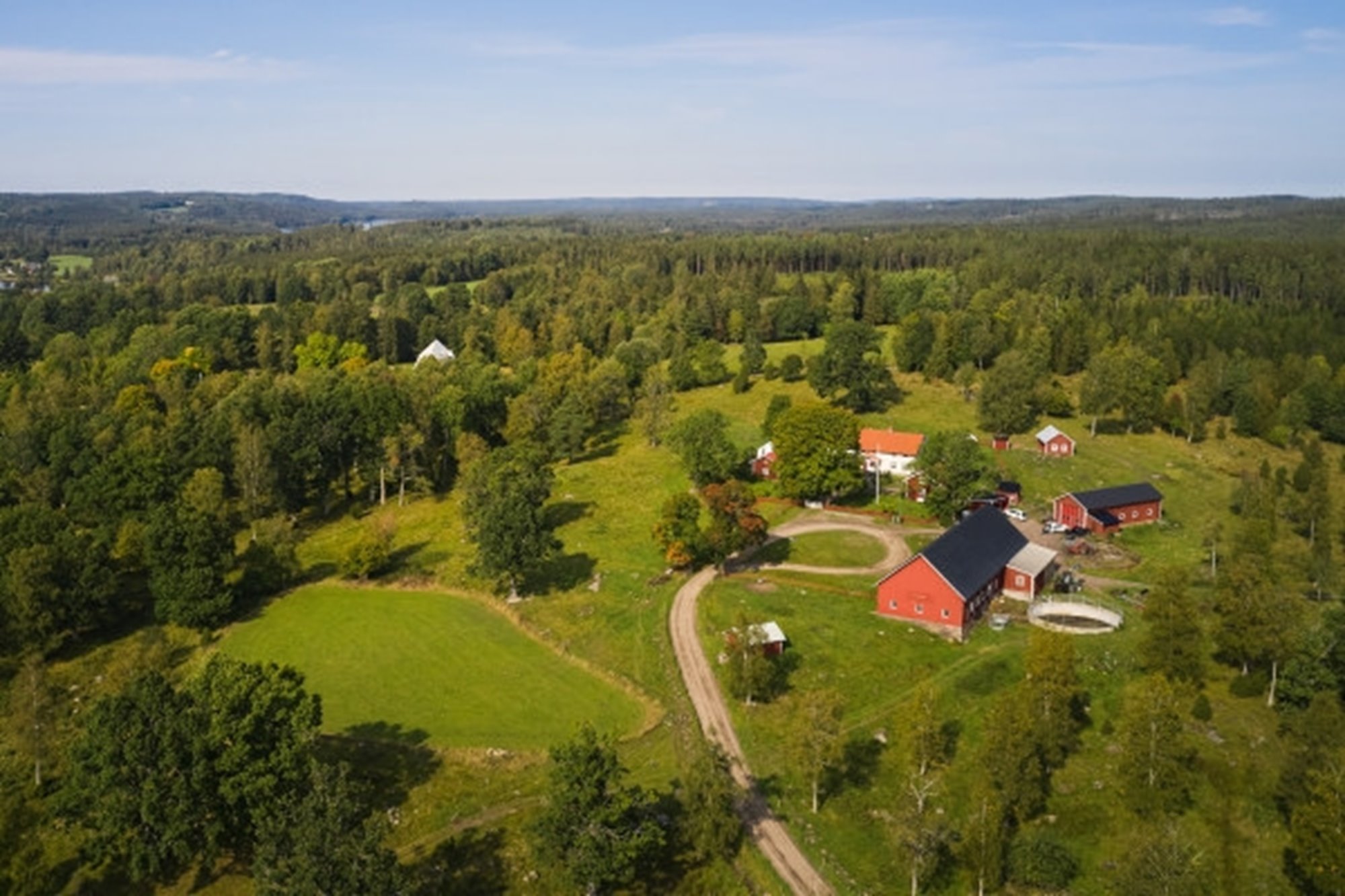 Skogsgård sedd från luften en sommardag med blå himmel. Vitt boningshus, röd ladugård och fält och blandskog runt omkring.. 